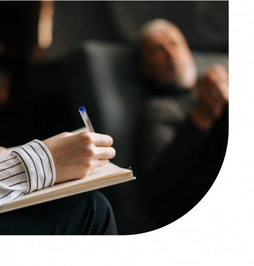 A person writing in a notebook with a person in a chair in the background